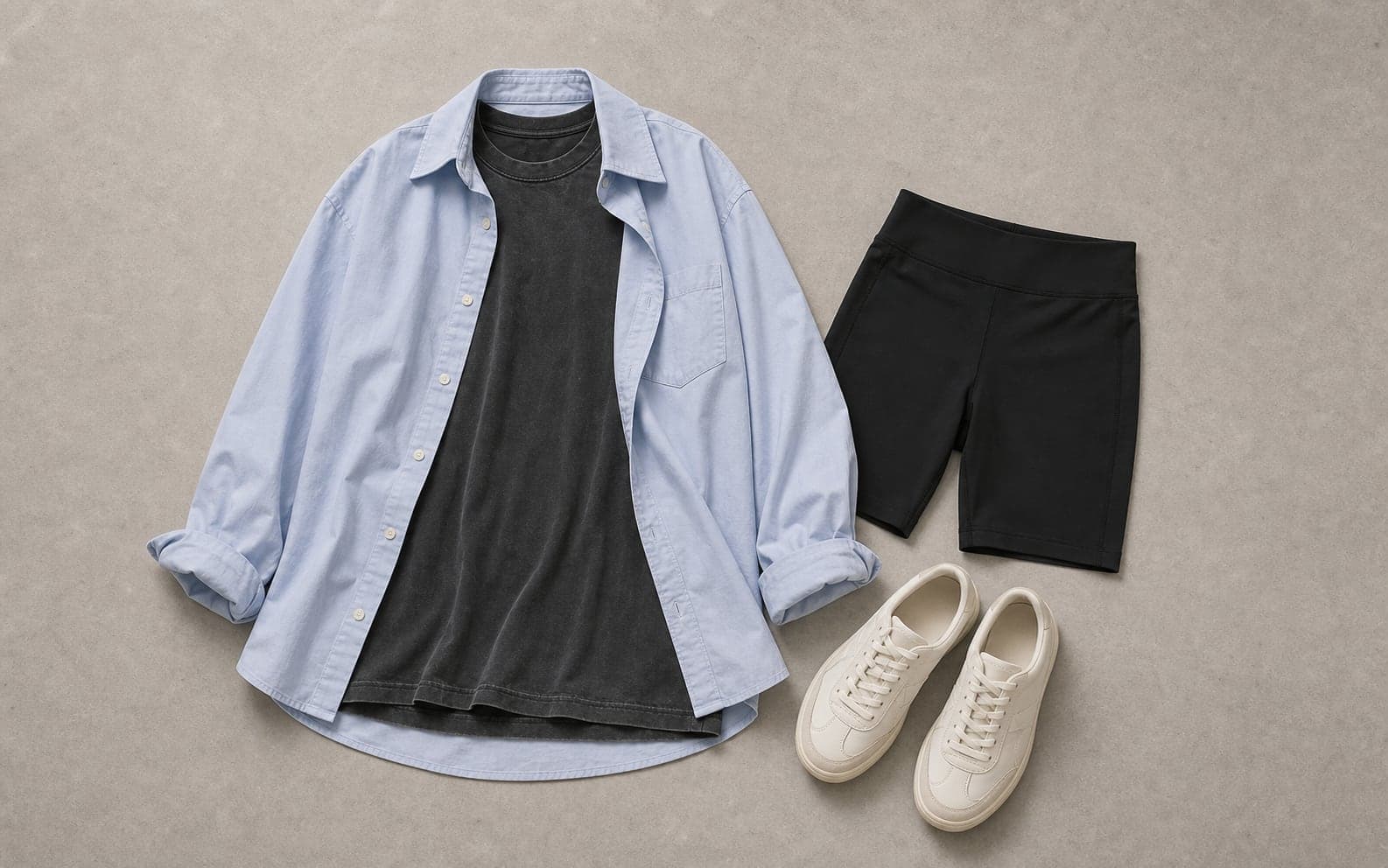 A complete casual outfit with an oversized charcoal tee, pale blue button-down shirt, black bike shorts, and cream sneakers.
