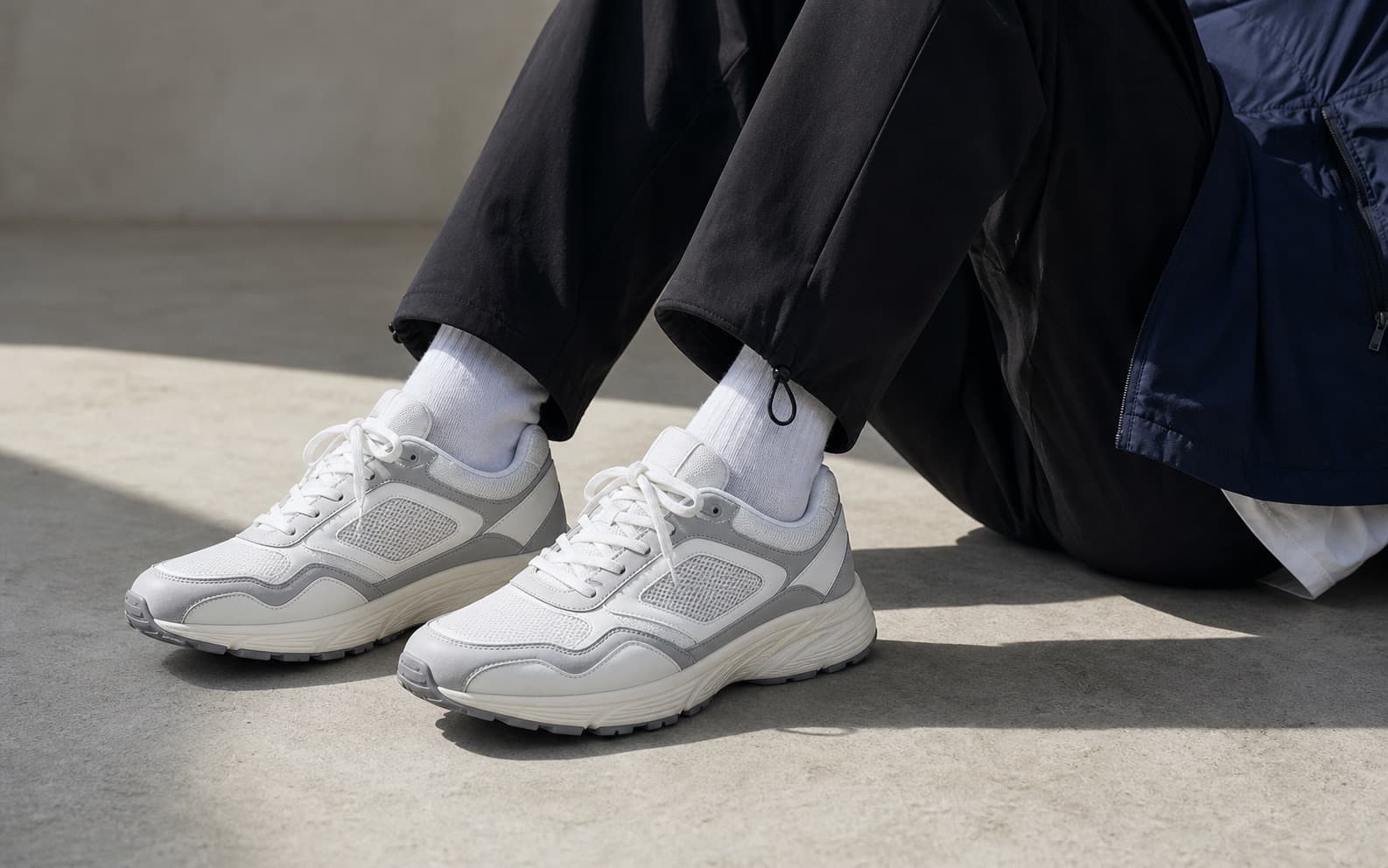 Clean white and gray training-inspired sneakers worn with straight dark pants.