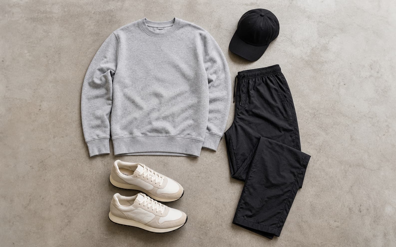 A gray crewneck sweatshirt, straight black nylon pants, cream retro runners, and black cap arranged as an outfit.