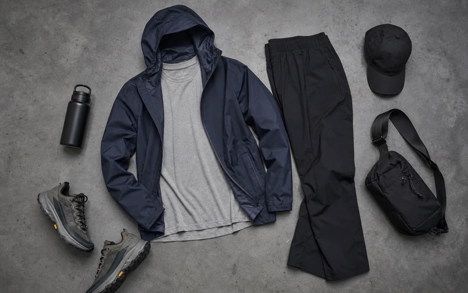 A tee, navy shell, black tech pants, trail runners, and technical accessories arranged as a weather outfit.