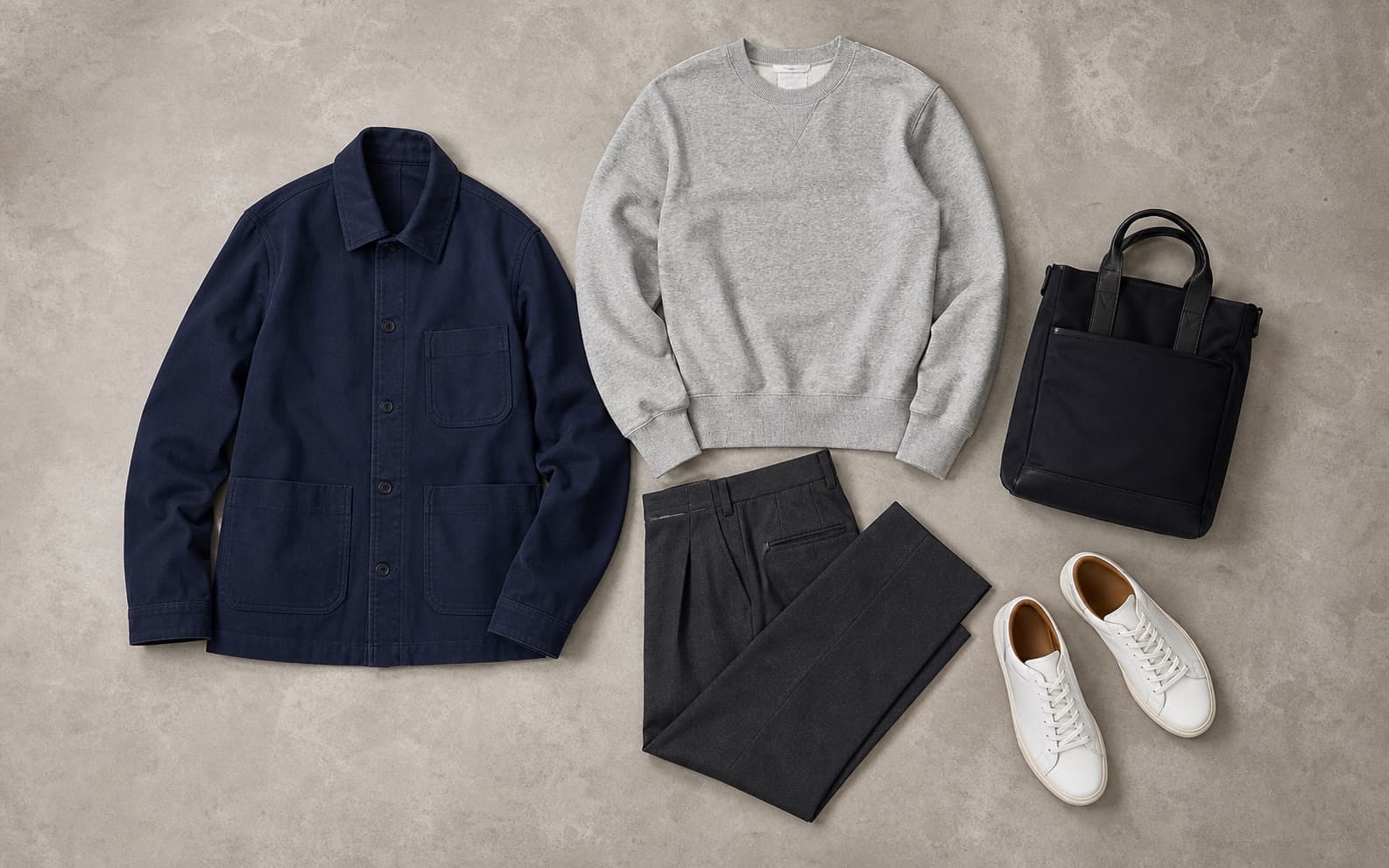 A blank gray crewneck, relaxed charcoal trousers, leather sneakers, and navy chore jacket arranged as a work-adjacent outfit.