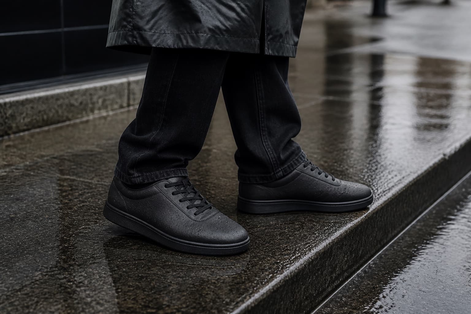 A close detail of black waterproof sneakers worn with dark denim on wet city pavement.