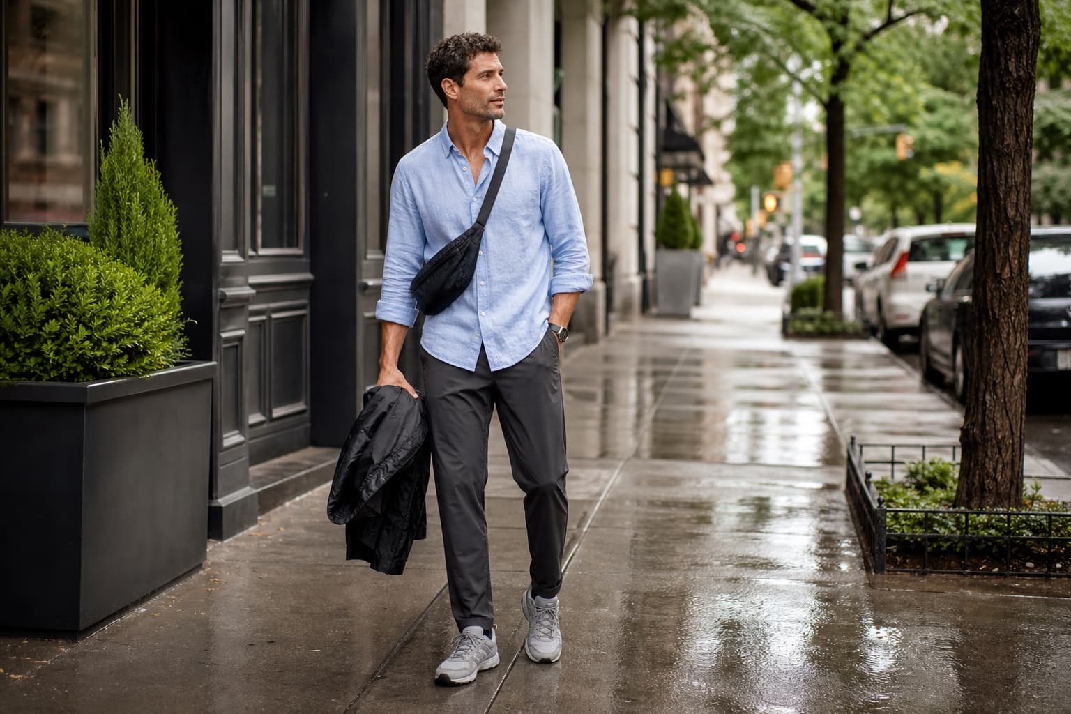 A man in a pale blue cotton-linen shirt, charcoal technical trousers, waterproof sneakers, crossbody bag, and packable shell.