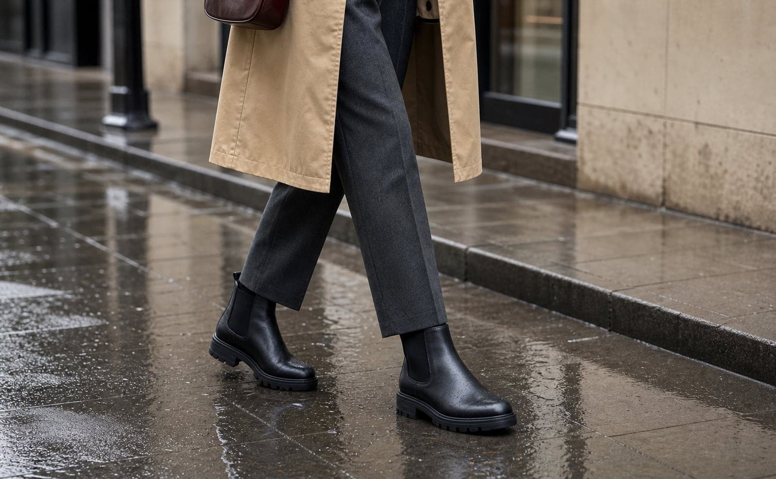 A close detail of black waterproof ankle boots worn with charcoal cropped trousers on wet pavement.