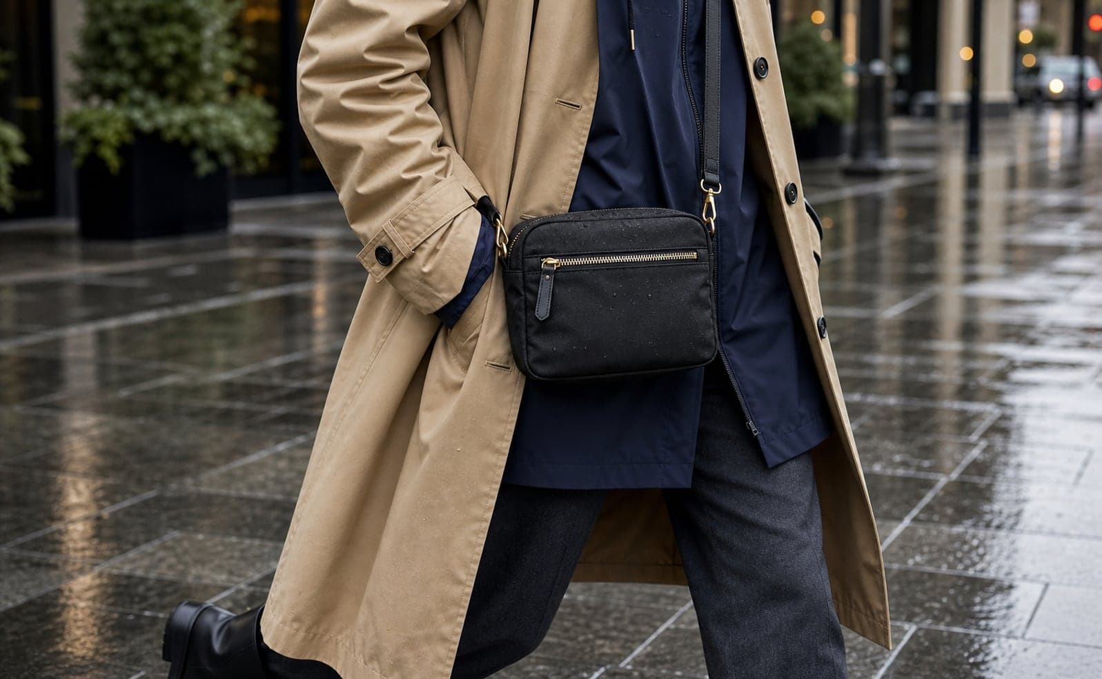 A close detail of a black weather-resistant crossbody bag worn over a navy rain shell on a rainy sidewalk.