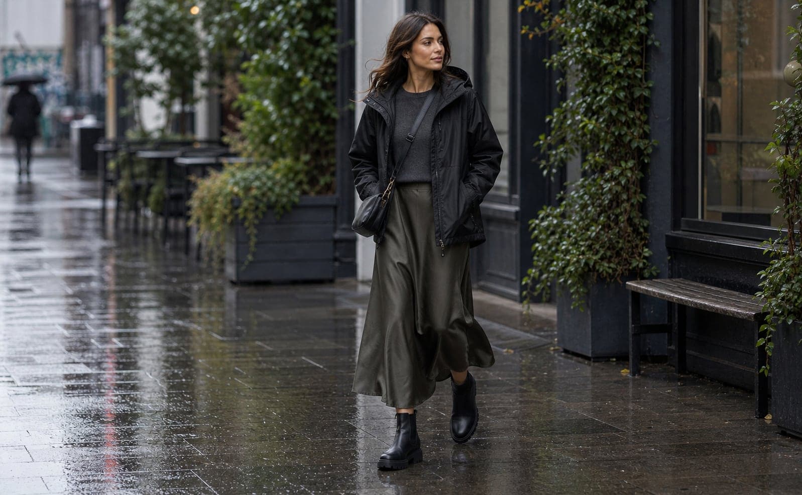 A woman wearing a black technical rain shell, charcoal knit top, dark satin midi skirt, lug-sole waterproof boots, and a crossbody bag on a rainy street.