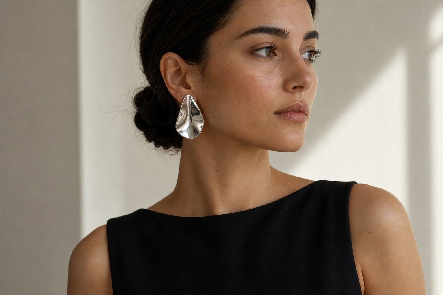 A close editorial portrait of a woman in a simple black top wearing polished silver sculptural earrings.