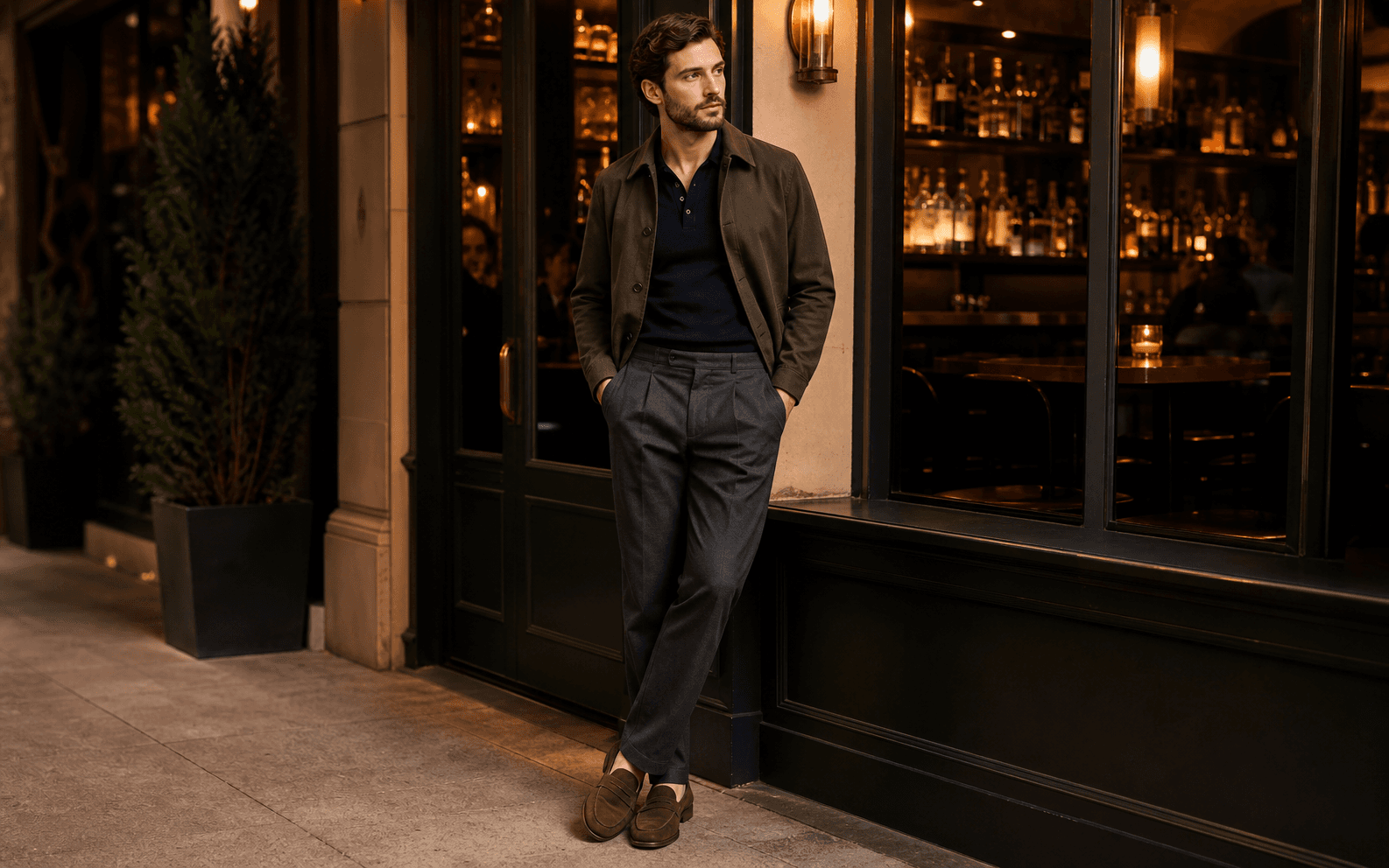 A man near a cocktail bar wearing a knit polo, relaxed trousers, loafers, and cropped jacket.