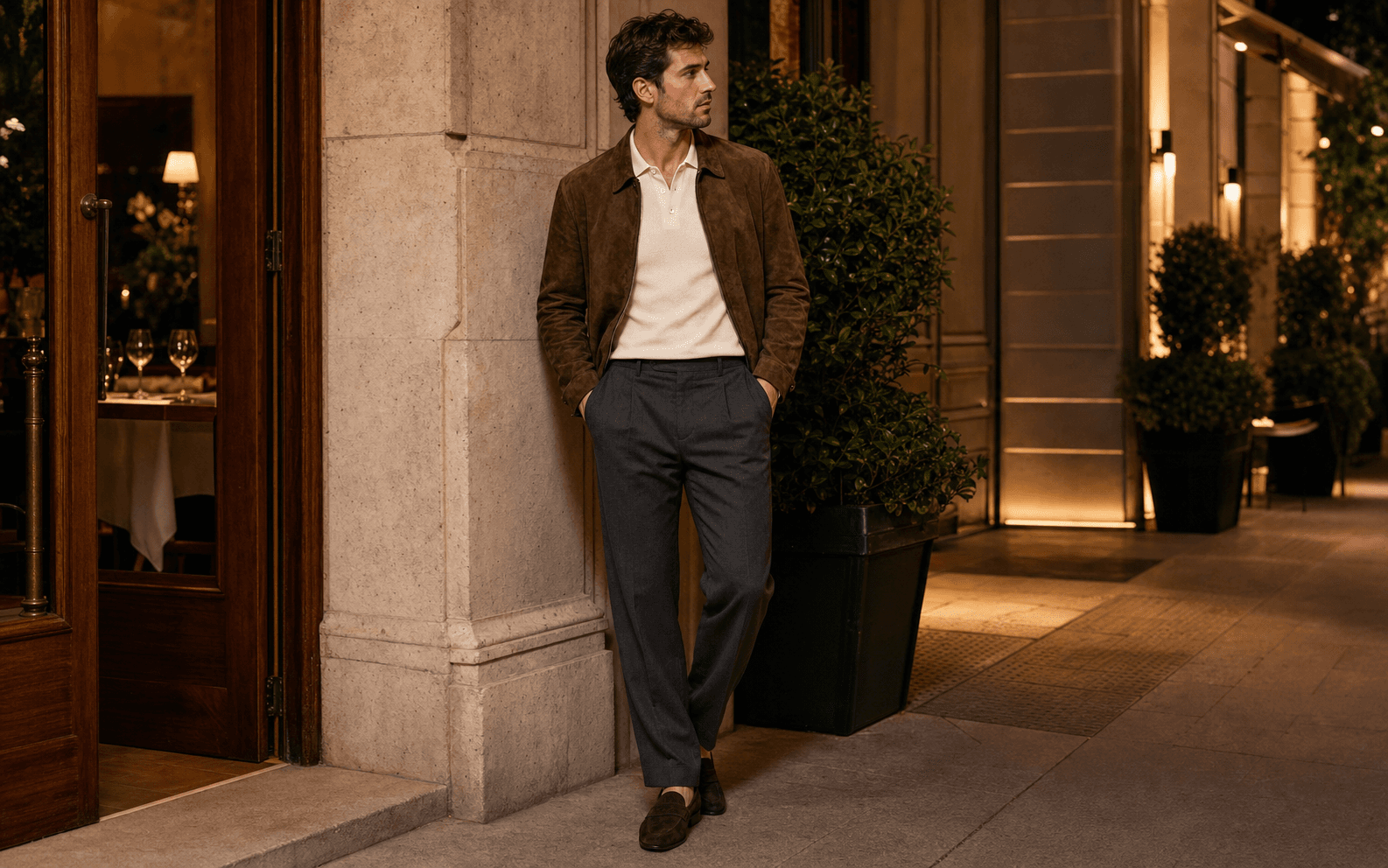 A man in a cream knit polo, charcoal trousers, chocolate suede jacket, and dark loafers in warm evening light.
