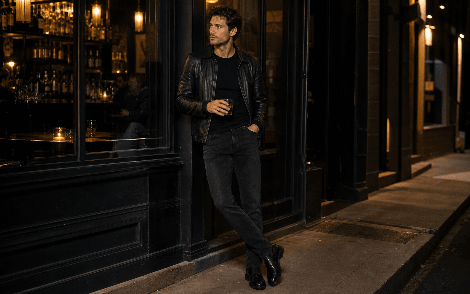 A man outside a low-lit bar wearing a black tee, washed black jeans, leather jacket, and black boots.