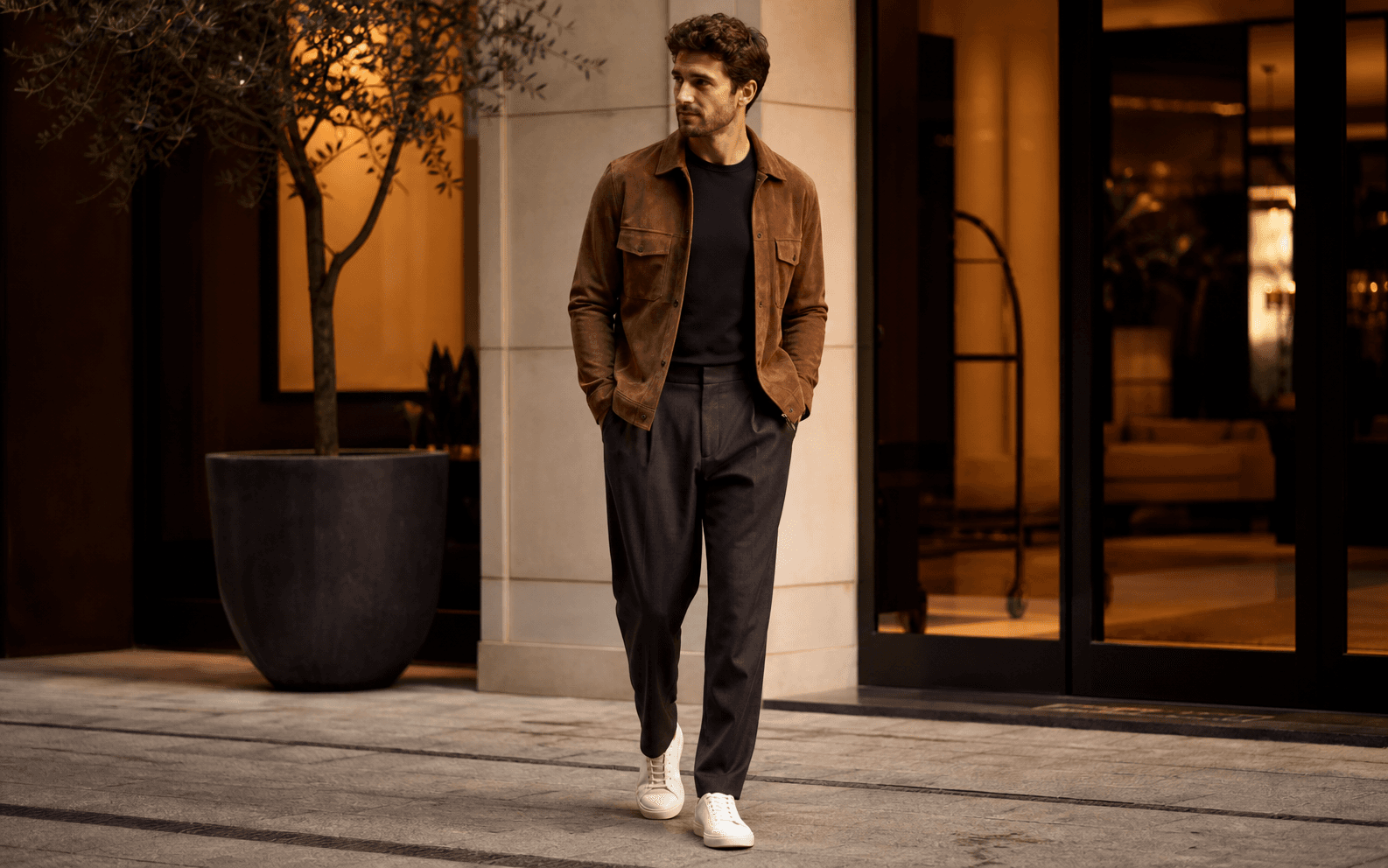 A man in a boutique hotel setting wearing a merino tee, relaxed trousers, suede overshirt, and minimal sneakers.
