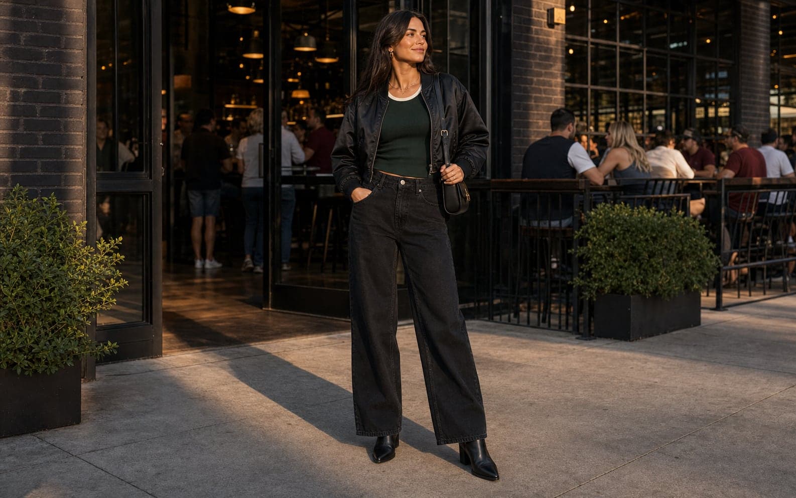 A woman outside a sports bar wearing a fitted forest green ribbed tank, cropped black bomber, relaxed black jeans, ankle boots, and a small bag.