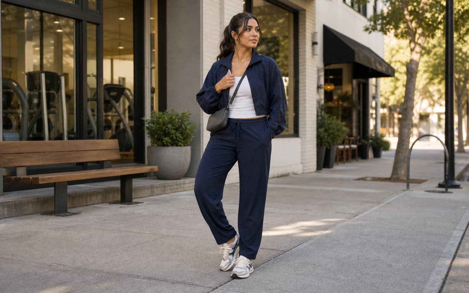 A woman in relaxed navy track pants, a fitted white ribbed tank, a cropped navy windbreaker, retro sneakers, and a compact crossbody bag standing outside a boutique gym and cafe.