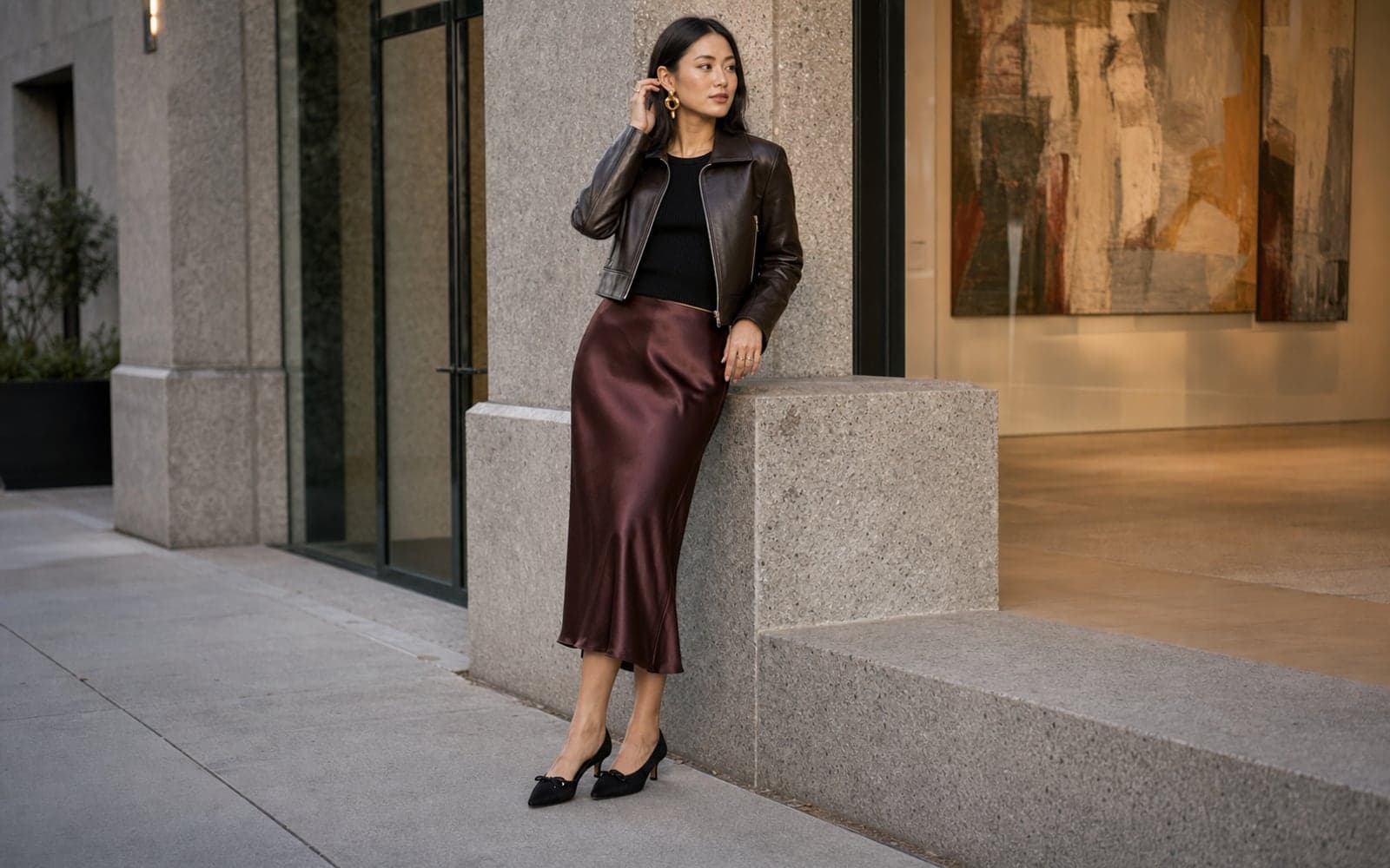 A woman wearing a burgundy satin midi skirt, black fitted knit, cropped leather jacket, and sculptural earrings leaning near a gallery entrance.