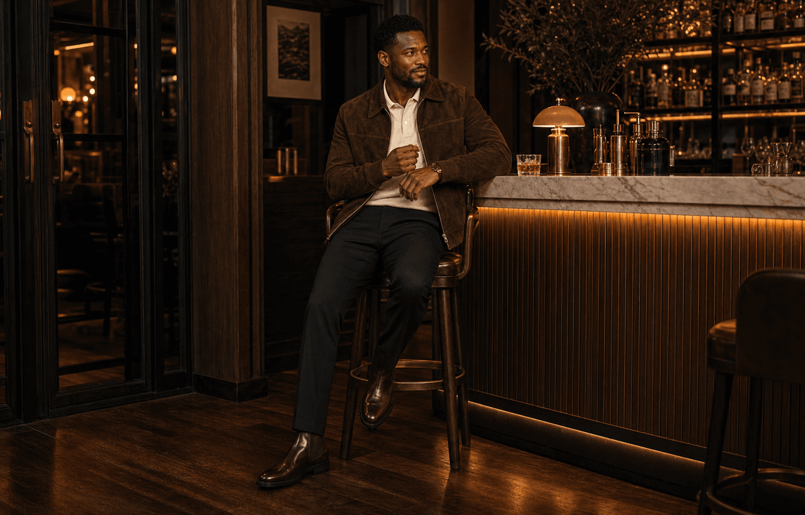 A man in a chocolate suede jacket, cream knit polo, dark straight jeans, and brown boots walking into a warmly lit restaurant at dusk.