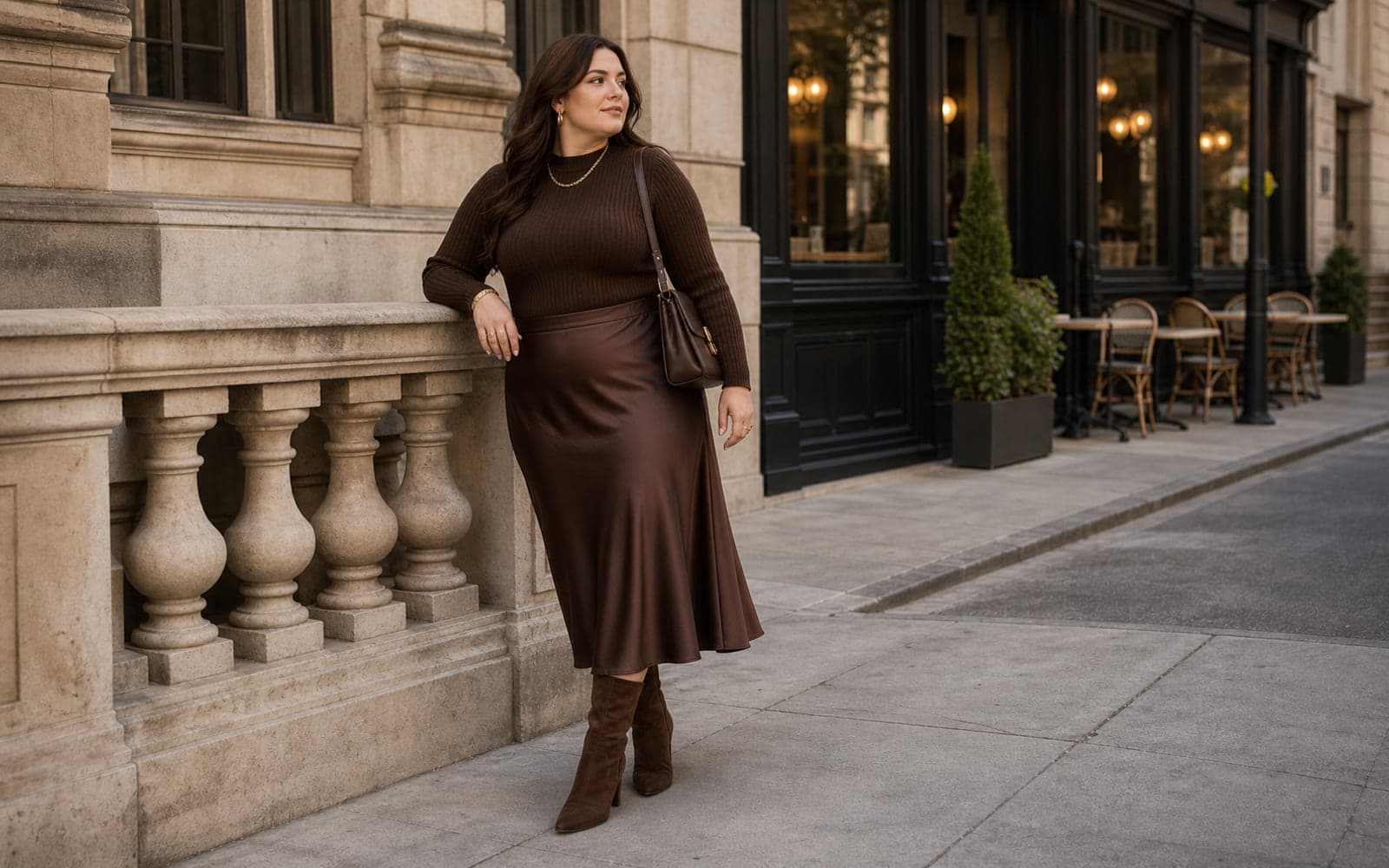 A woman in a tonal chocolate ribbed knit top, satin midi skirt, brown suede boots, gold jewelry, and a deep brown shoulder bag leaning against a stone railing.