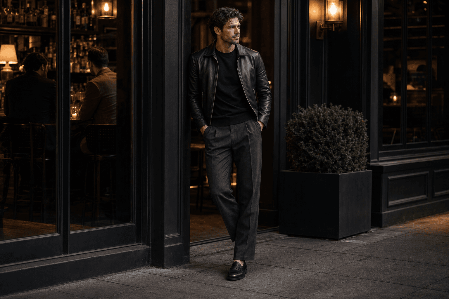 A man outside a cocktail bar wearing a black tee, charcoal trousers, black loafers, and a cropped leather jacket.