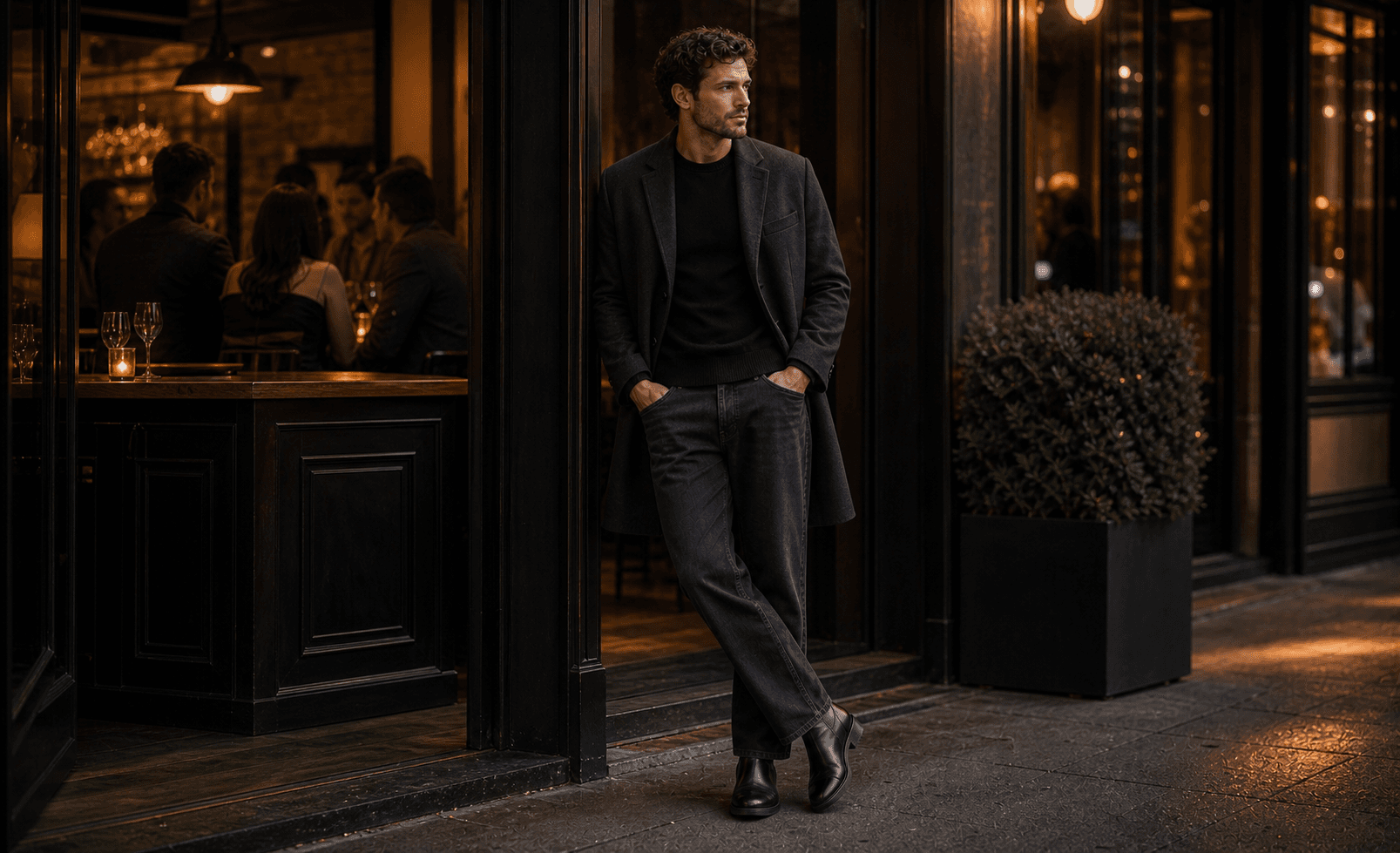 A man outside a warmly lit date-drinks spot wearing a black sweater, dark jeans, leather boots, and a polished outer layer.