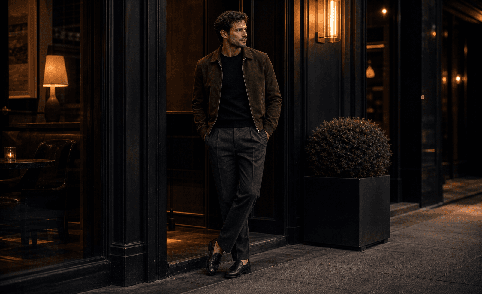 A man at an evening lounge entrance wearing a black knit tee, charcoal trousers, suede bomber, and dark loafers.