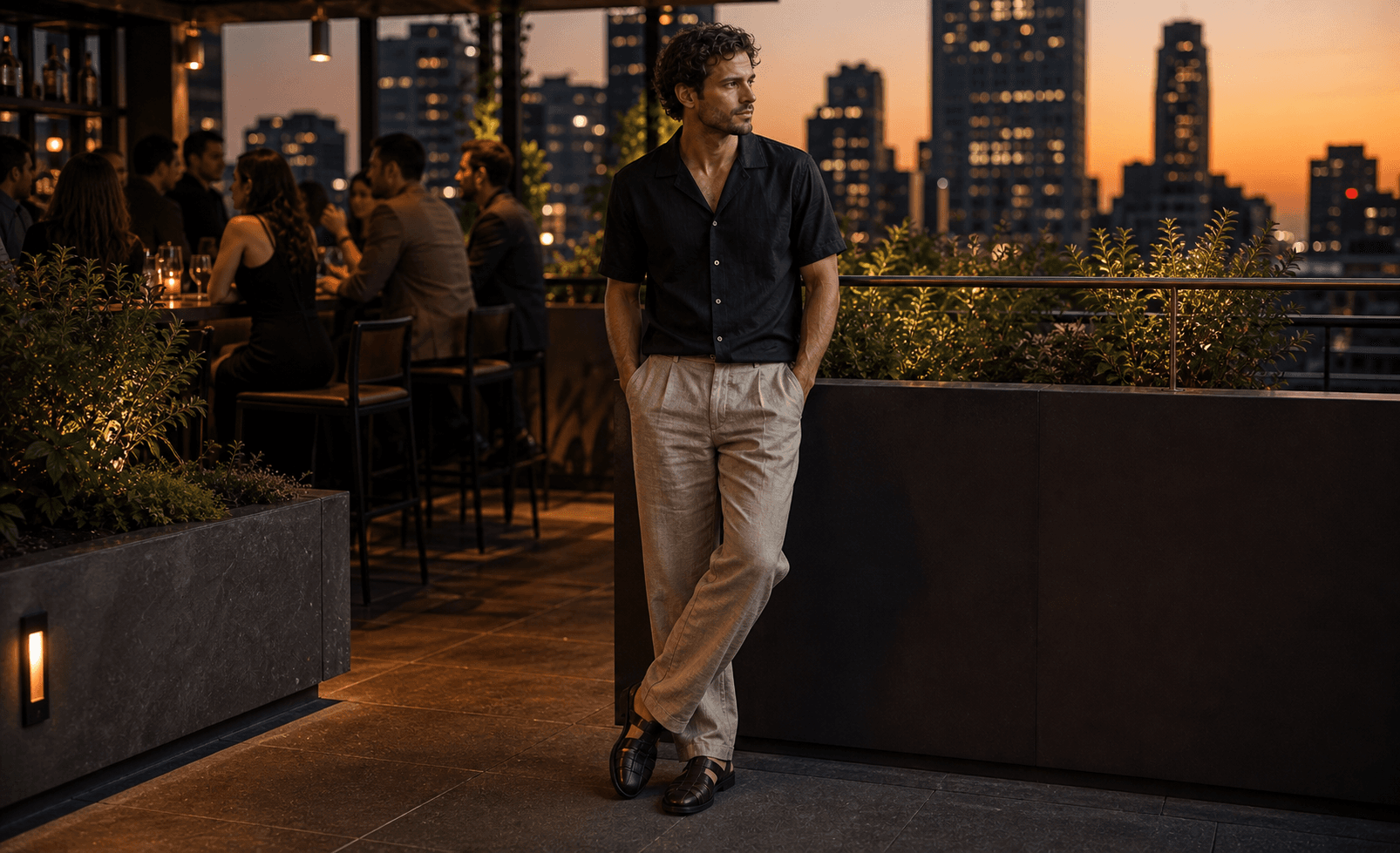 A man on a summer city patio wearing a dark camp collar shirt, relaxed light trousers, and dark leather footwear.