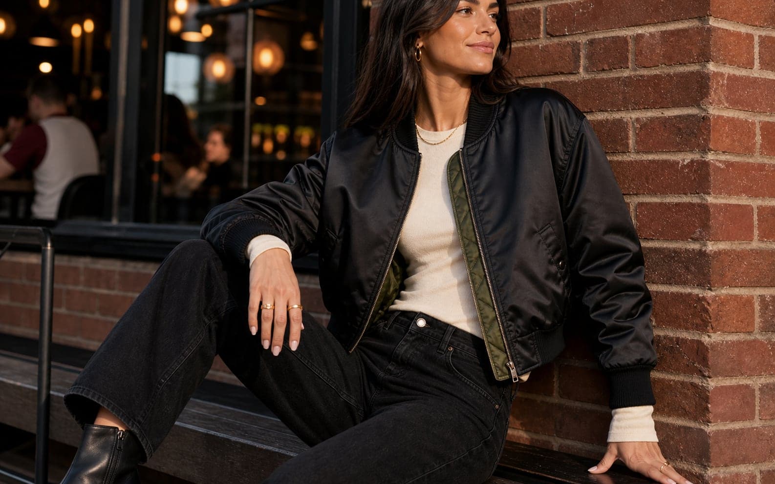 A woman wearing a cropped black bomber with a cream top and dark wide-leg jeans at night.