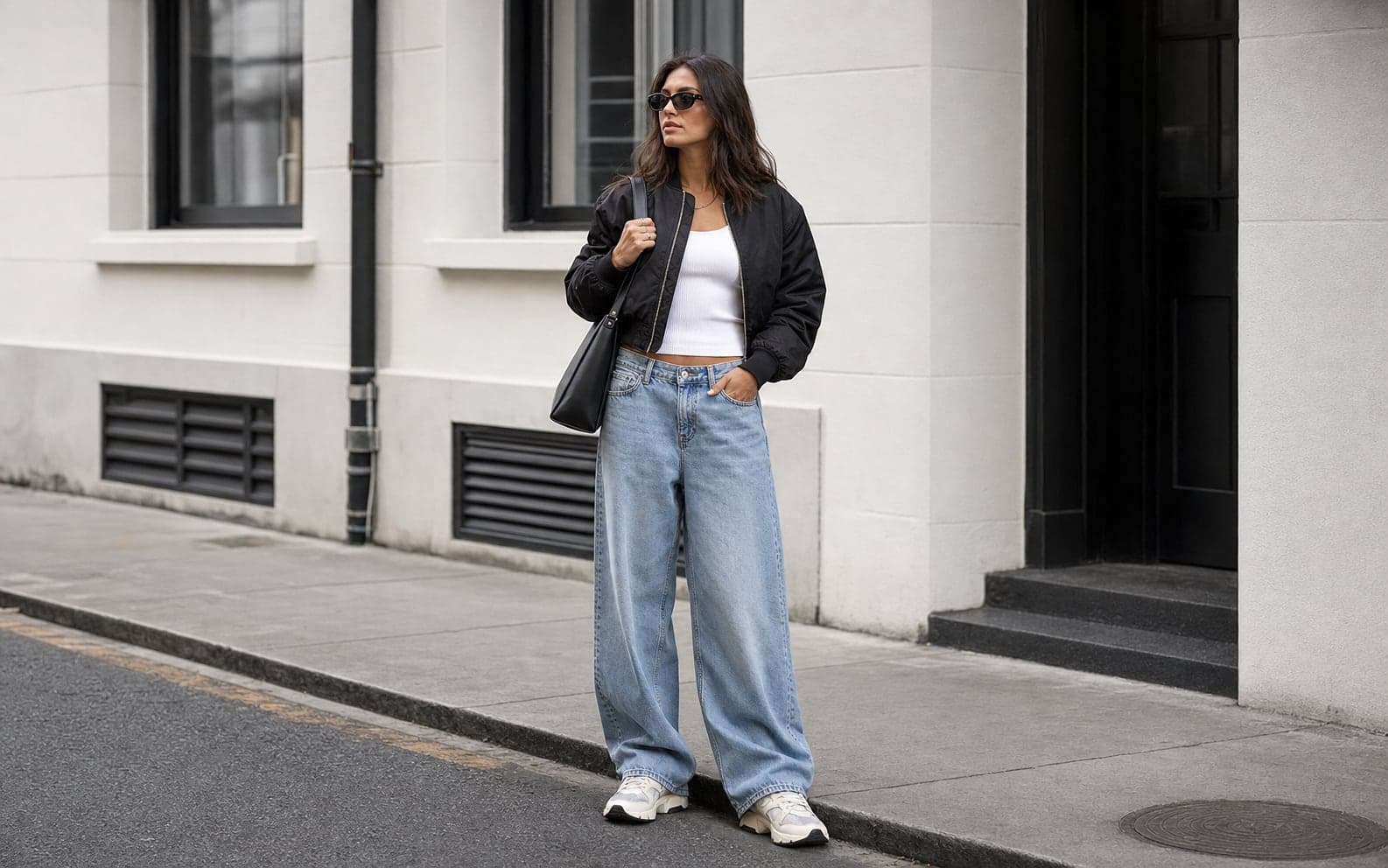 A woman in relaxed wide-leg denim, a fitted white tank, a cropped black bomber, substantial sneakers, and a structured shoulder bag.