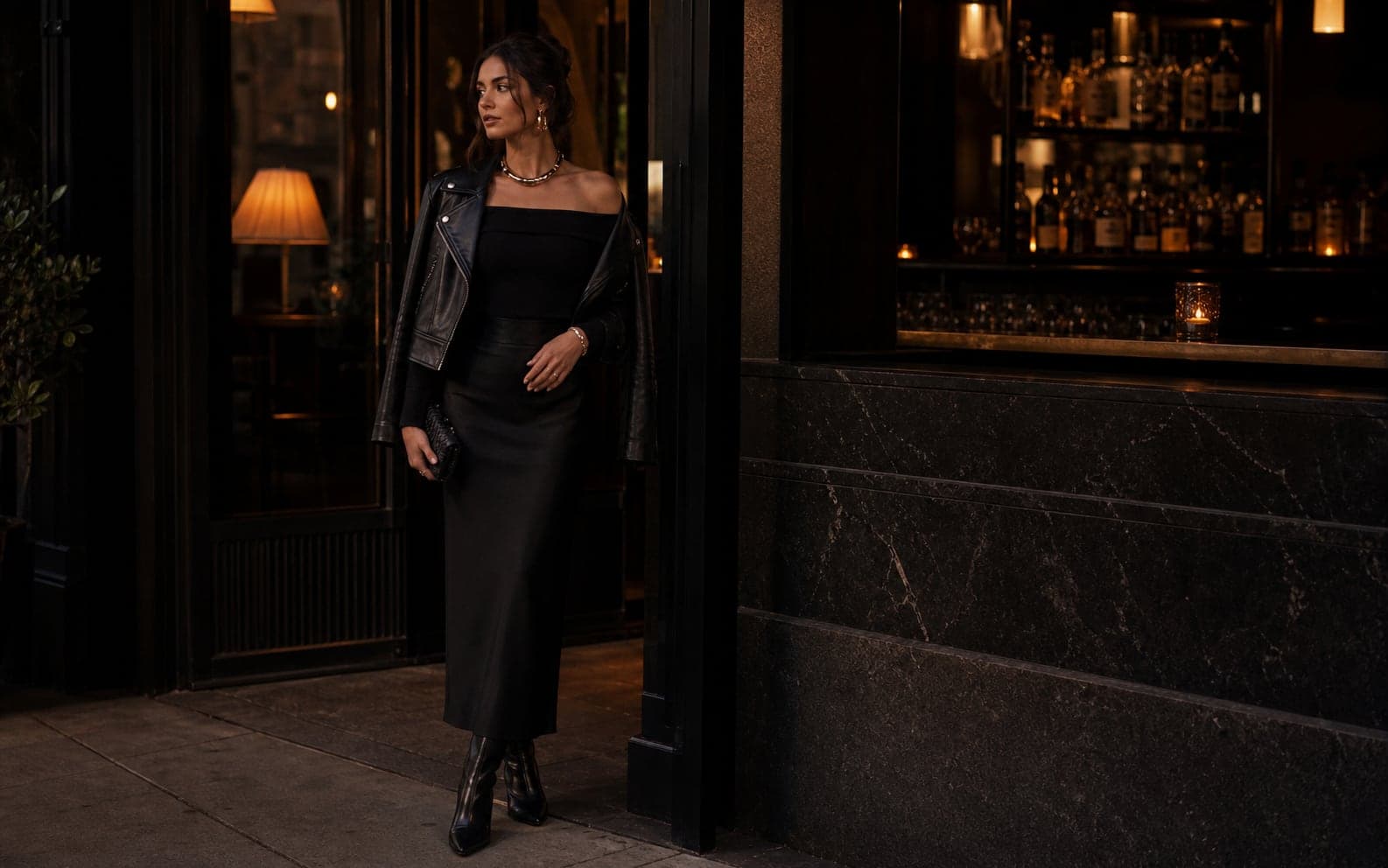 A woman near a low-lit bar wearing a black column skirt, fitted off-shoulder top, leather jacket, heeled boots, and metallic jewelry.