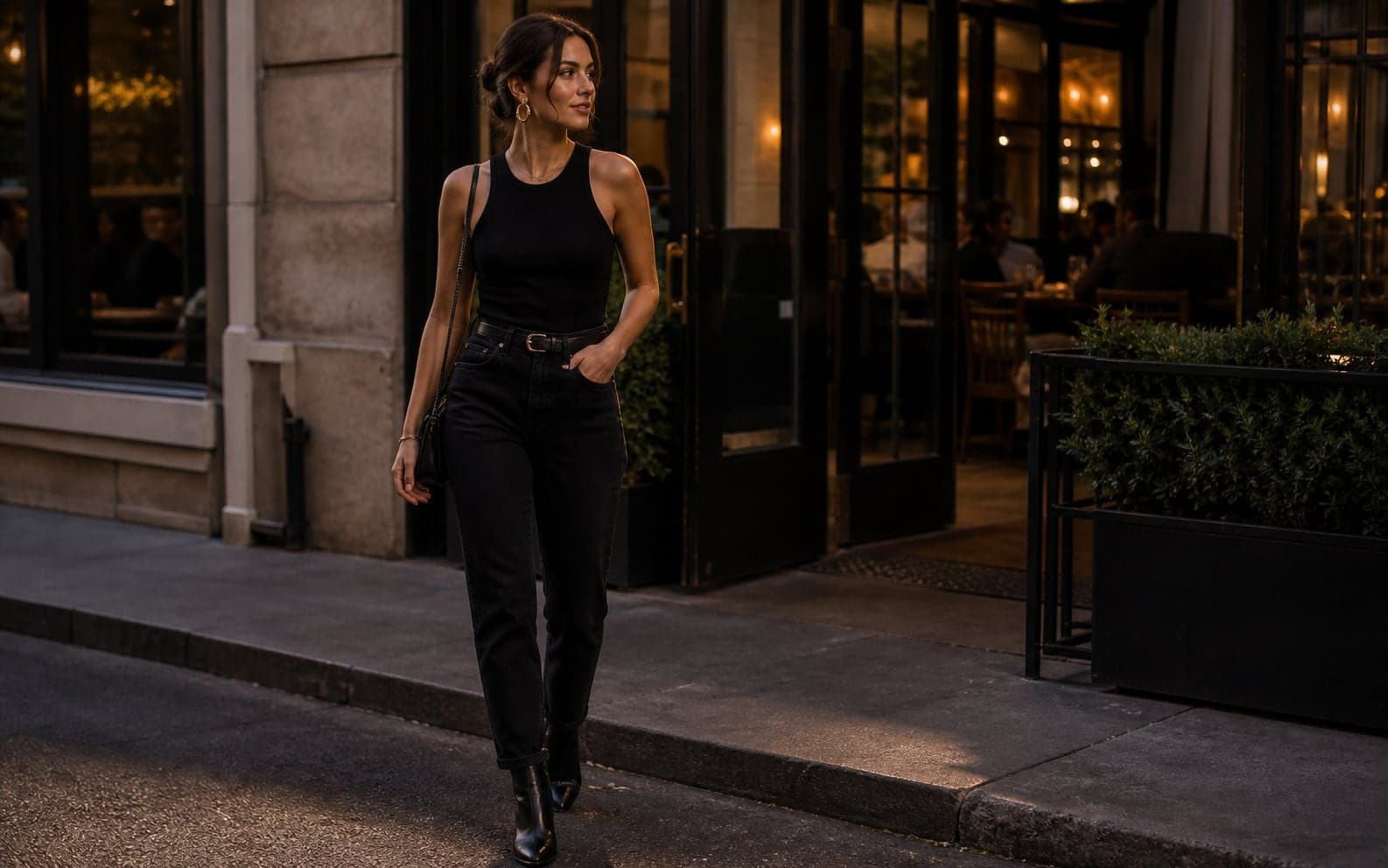 A woman outside a casual restaurant wearing a black tank, black trousers, belt, boots, and bold earrings.