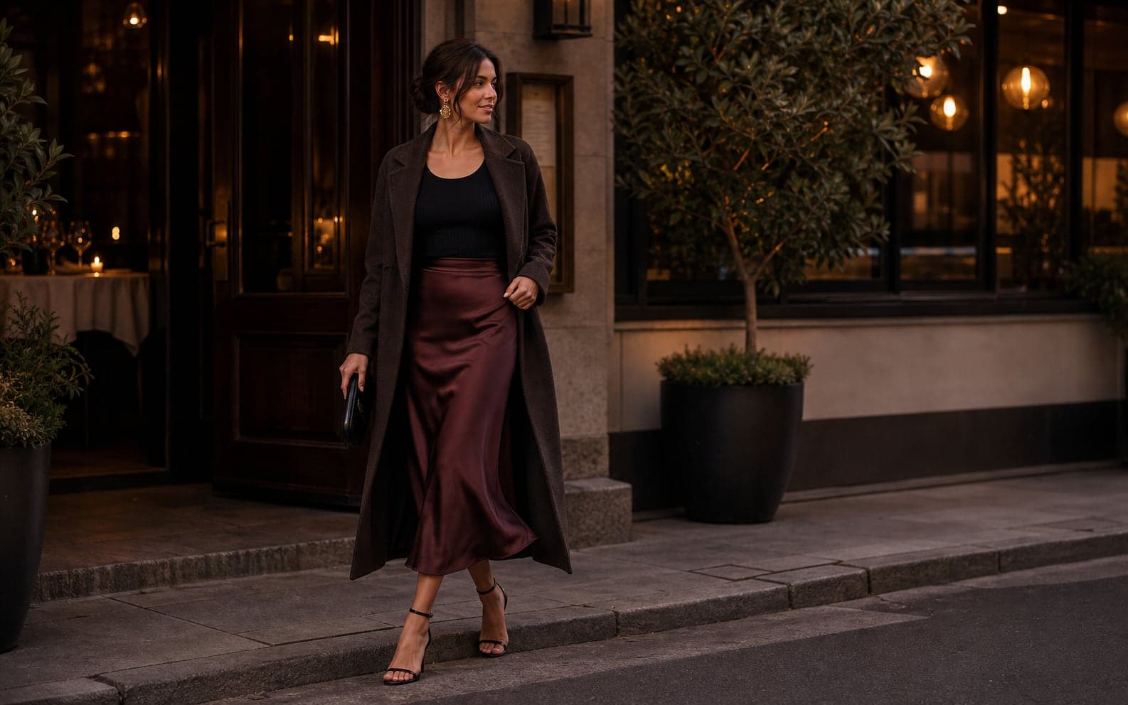A woman outside a warmly lit restaurant wearing a satin midi skirt, fitted knit top, long coat, heeled sandals, and sculptural earrings.