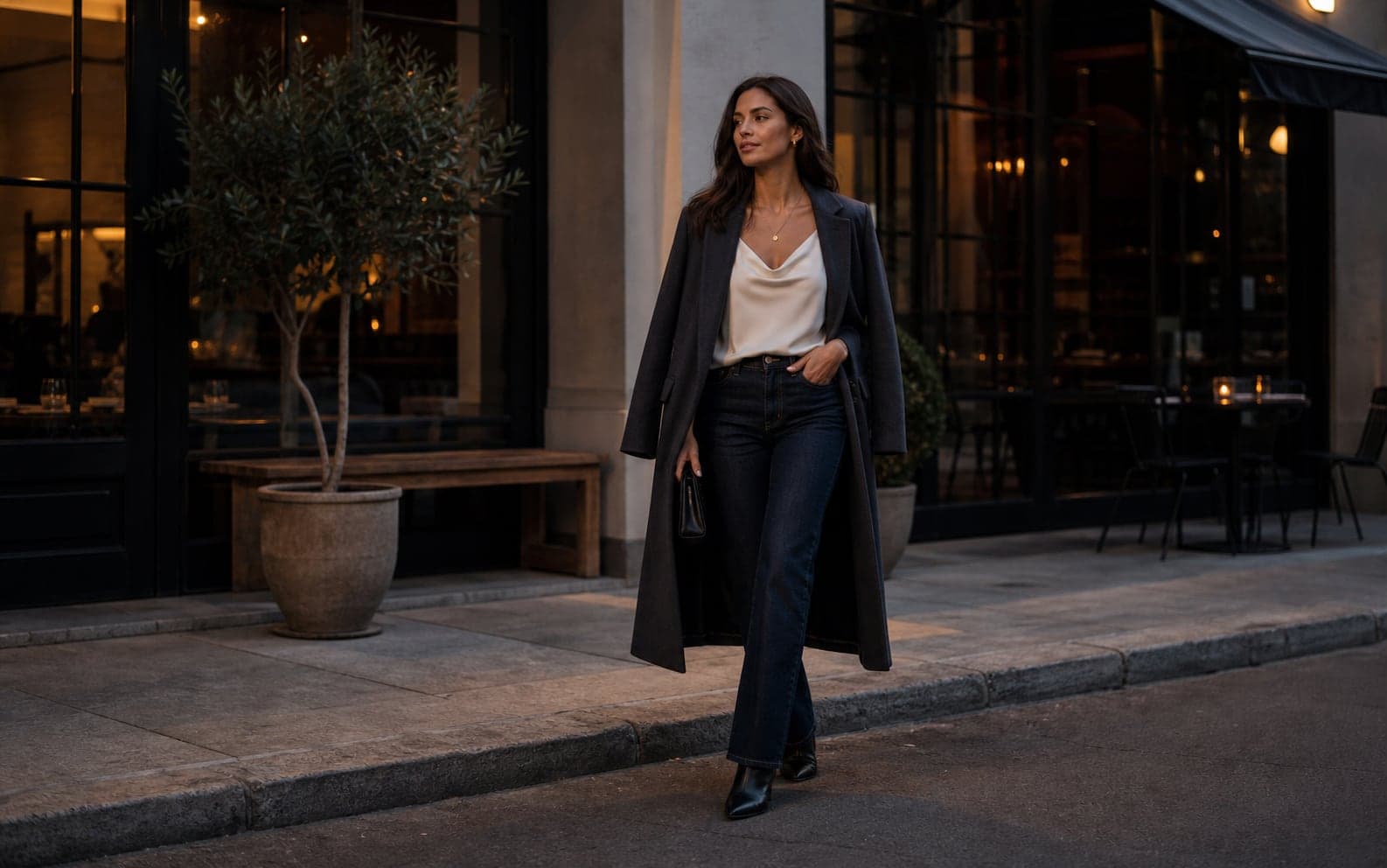 A woman outside an upscale casual restaurant wearing dark denim, a satin top, long coat, and pointed footwear.
