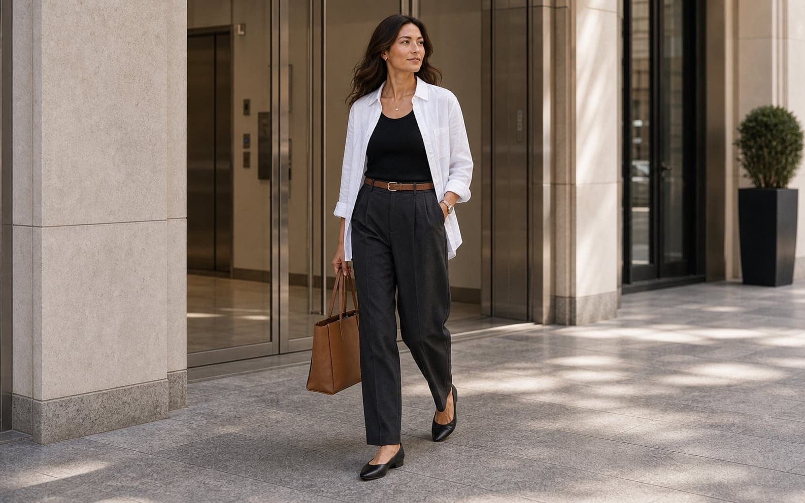 A woman wearing a white button-up over a black tank with charcoal trousers, clean flats, and structured bag.