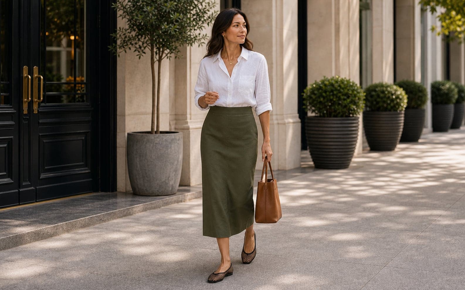 A woman wearing a white linen button-up, olive column skirt, mesh flats, and structured tan bag.